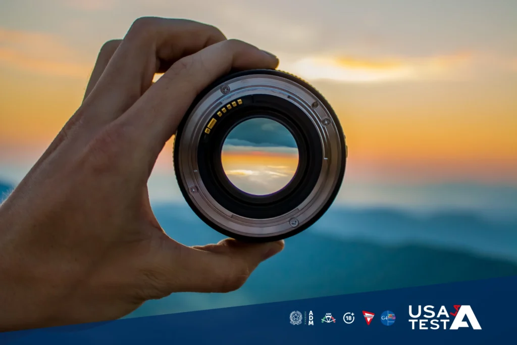 Mano che tiene un obiettivo fotografico puntato verso un paesaggio al tramonto, con vista sfocata sulle montagne e loghi del progetto Usa la Testa nella parte inferiore dell’immagine.